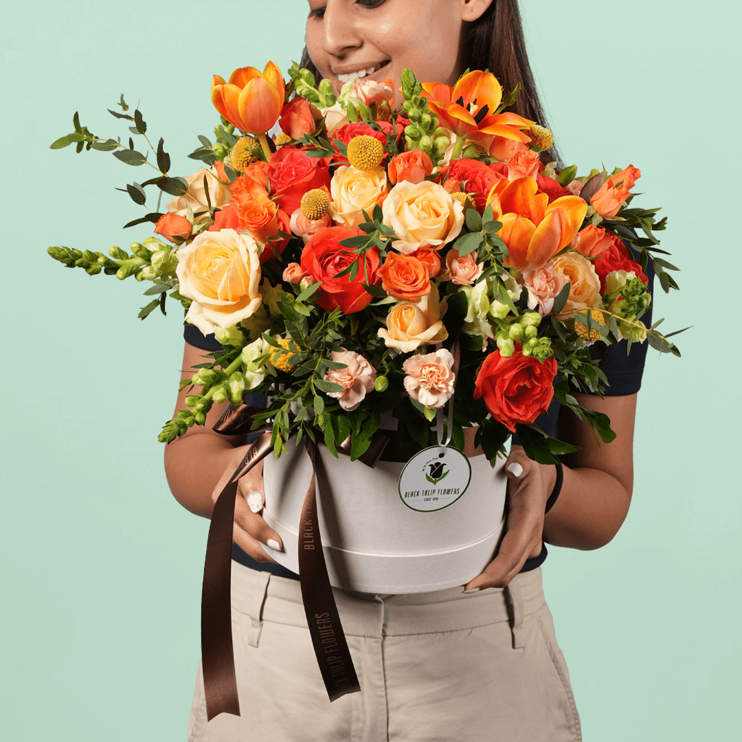 A white flower box filled with orange Tulips, Roses, Snapdragons, Craspedia, Spray Carnations and Eucalyptus