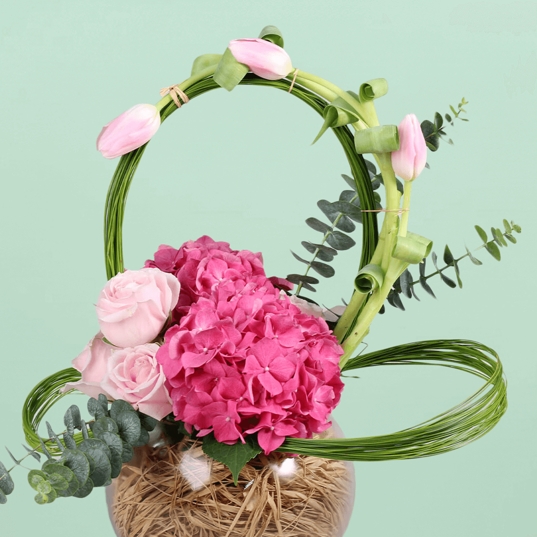 A glass fishbowl arrangement with assorted pink tulips, hydrangea and roses with looped steel grass.