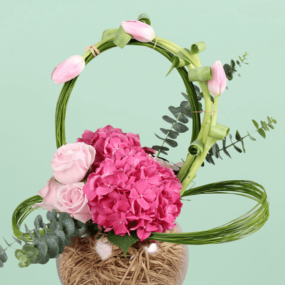 A glass fishbowl arrangement with assorted pink tulips, hydrangea and roses with looped steel grass.
