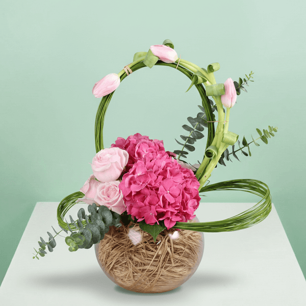 A glass fishbowl arrangement with assorted pink tulips, hydrangea and roses with looped steel grass.