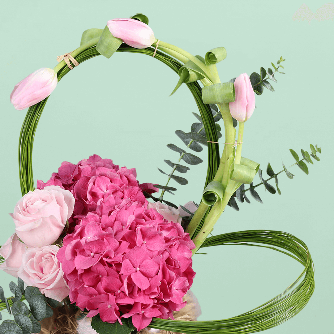 A glass fishbowl arrangement with assorted pink tulips, hydrangea and roses with looped steel grass.