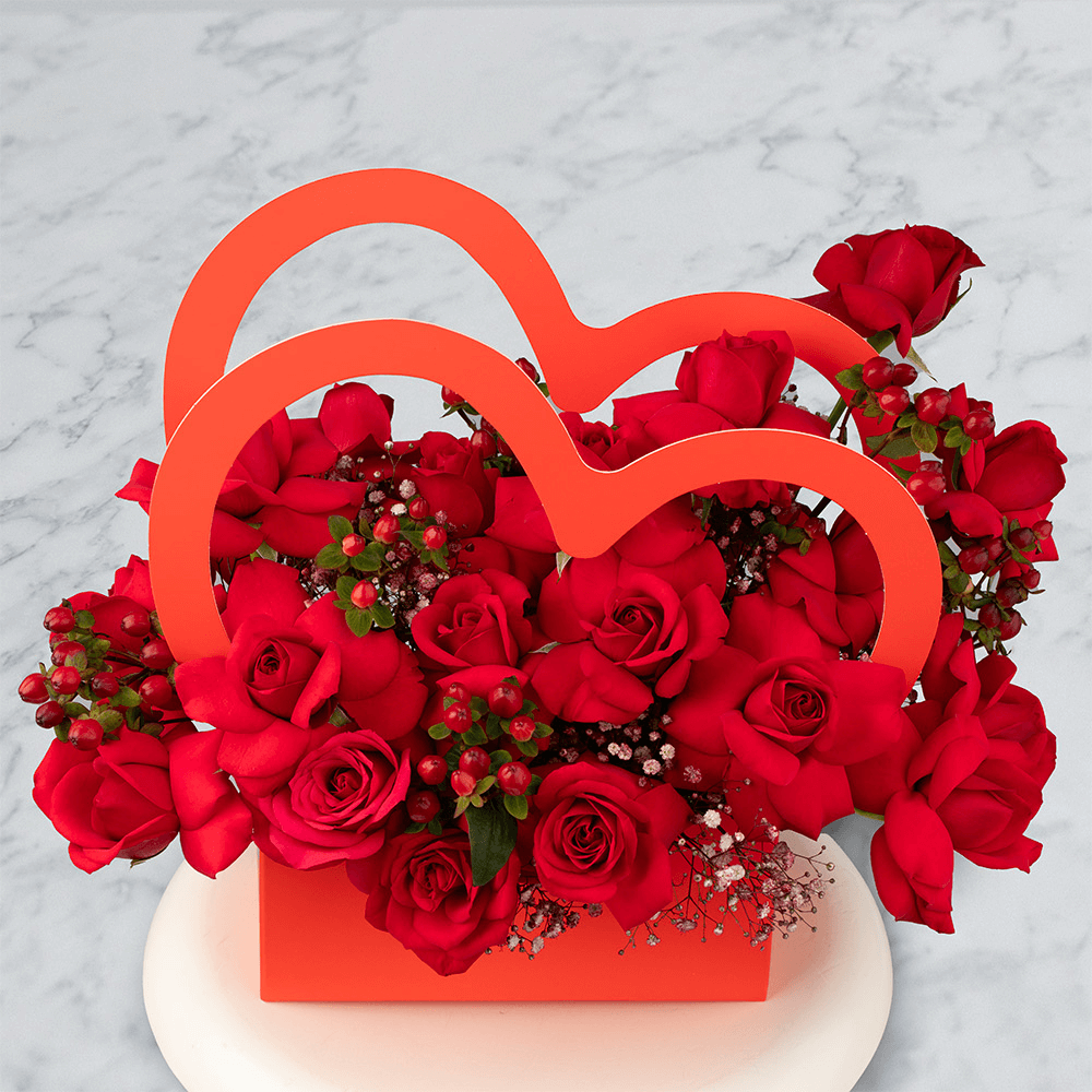 Red Roses with berries and Gypsophila arranged in a red heart-shaped flower box.
