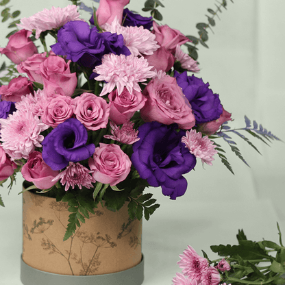 Purple Eustoma, pink Roses and Chrysanthemums with greenery arranged in a round brown flower box.