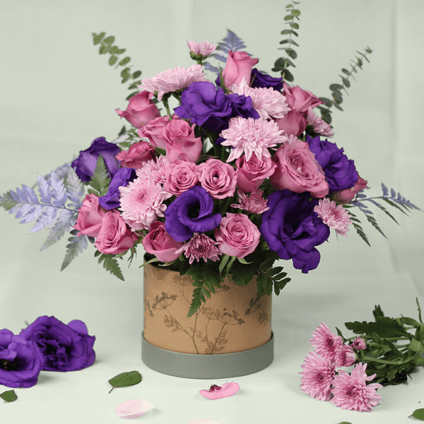 Purple Eustoma, pink Roses and Chrysanthemums with greenery arranged in a round brown flower box.