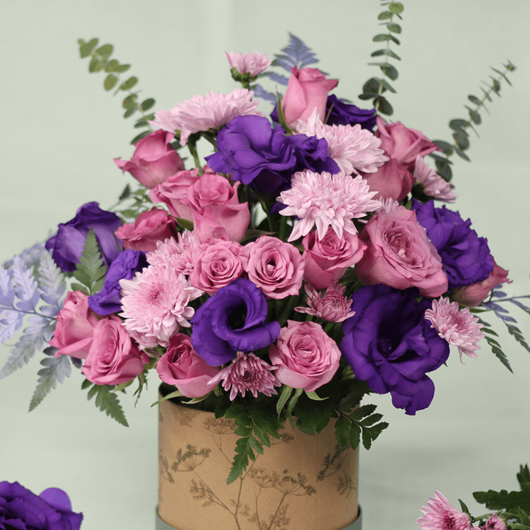 Purple Eustoma, pink Roses and Chrysanthemums with greenery arranged in a round brown flower box.