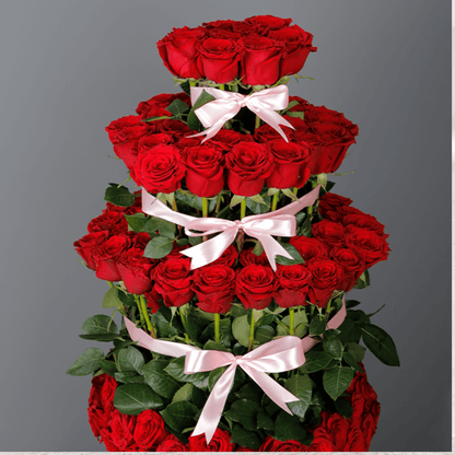 Tiered arrangement featuring red roses in a pink round box, finished with soft pink ribbon bows.