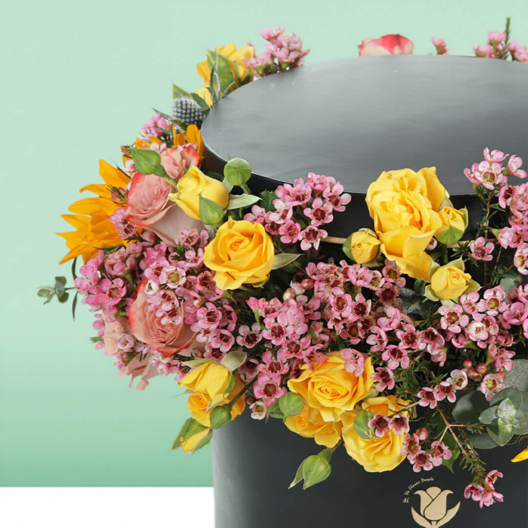 Sunflowers, yellow Spray Roses and pink Wax Flowers arranged around a black round flower box.