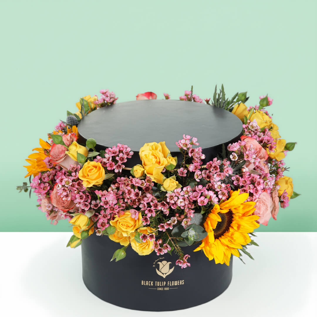 Sunflowers, yellow Spray Roses and pink Wax Flowers arranged around a black round flower box.