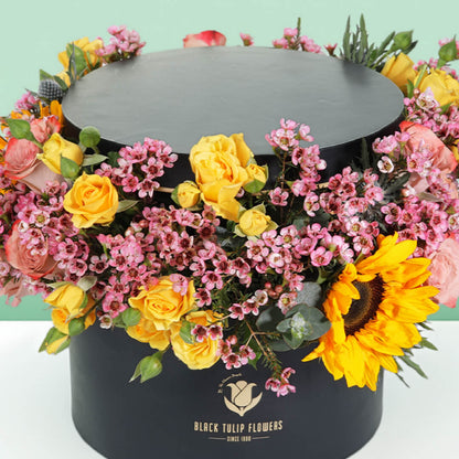 Sunflowers, yellow Spray Roses and pink Wax Flowers arranged around a black round flower box.