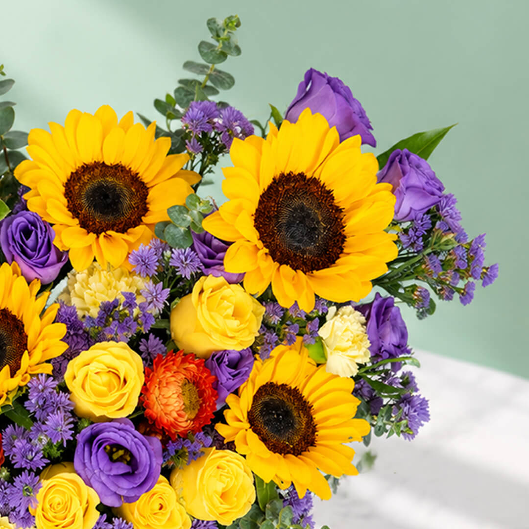Sunflower, rose and blue daisy flower arrangement with statice, helichrysum, eustoma and eucalyptus in a round black box.