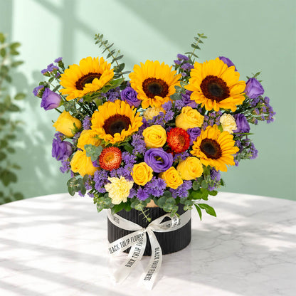 Sunflower, rose and blue daisy flower arrangement with statice, helichrysum, eustoma and eucalyptus in a round black box.