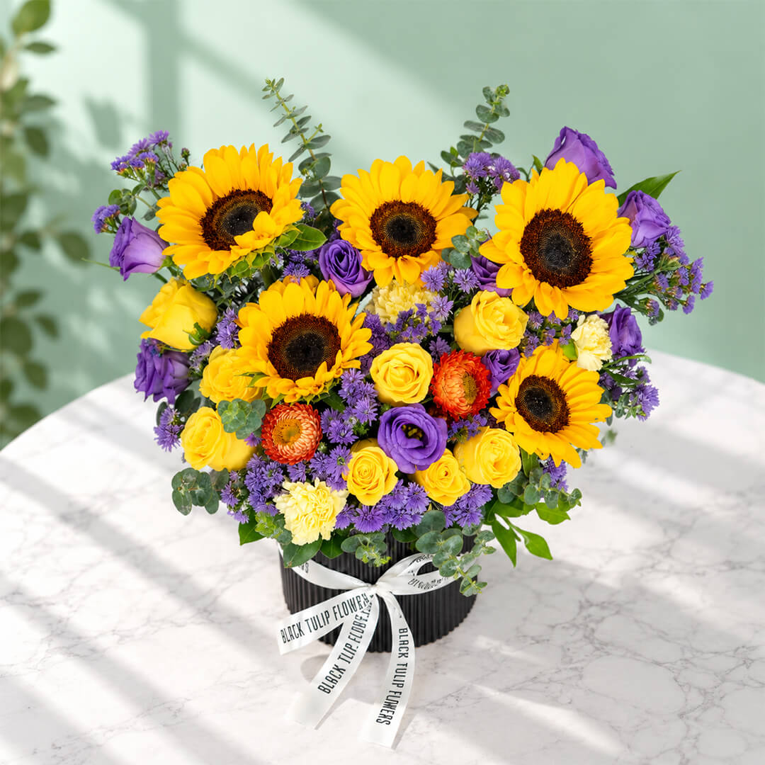 Sunflower, rose and blue daisy flower arrangement with statice, helichrysum, eustoma and eucalyptus in a round black box.