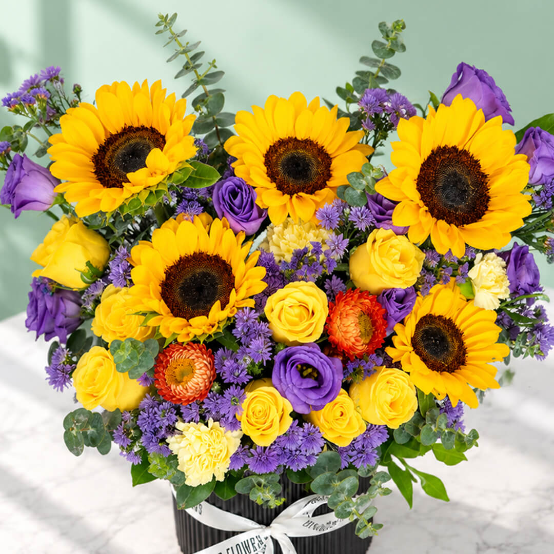 Sunflower, rose and blue daisy flower arrangement with statice, helichrysum, eustoma and eucalyptus in a round black box.