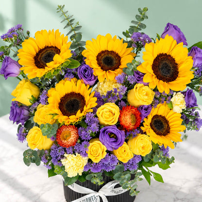 Sunflower, rose and blue daisy flower arrangement with statice, helichrysum, eustoma and eucalyptus in a round black box.