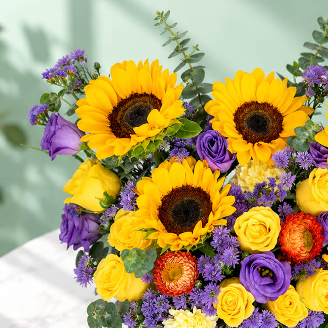 Sunflower, rose and blue daisy flower arrangement with statice, helichrysum, eustoma and eucalyptus in a round black box.
