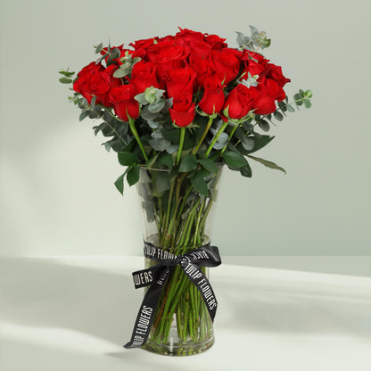 25 fresh red roses with greenery arranged in a clear hourglass glass vase