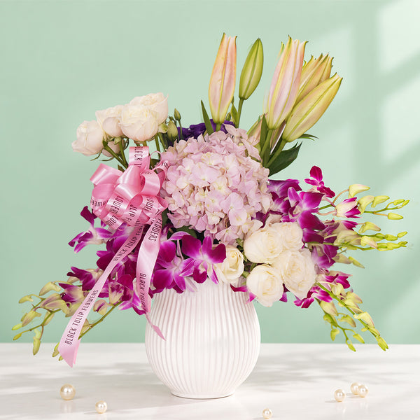Elegant pastel flower arrangement with pink hydrangea, white roses, lilies, and purple orchids in a white ribbed vase with a pink ribbon.