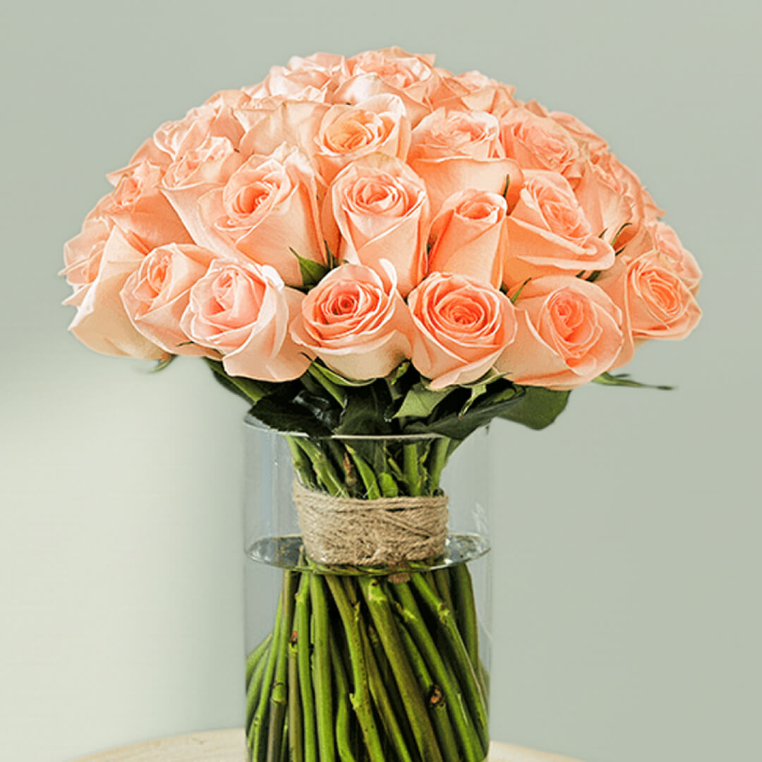 30 handpicked peach roses with rich green foliage in a modern transparent glass vase.