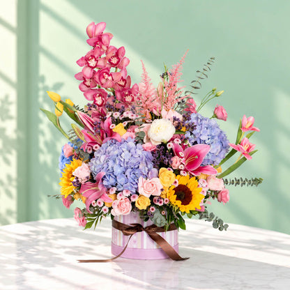 Pink round flower box featuring 50–60 stems of assorted hydrangeas, peonies, sunflowers, tulips, roses, lilies, cymbidium orchids and eucalyptus greenery.