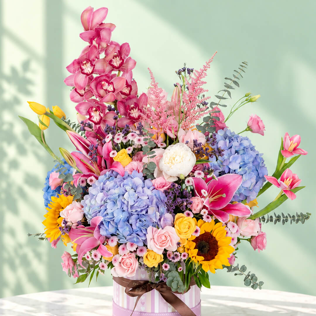 Pink round flower box featuring 50–60 stems of assorted hydrangeas, peonies, sunflowers, tulips, roses, lilies, cymbidium orchids and eucalyptus greenery.
