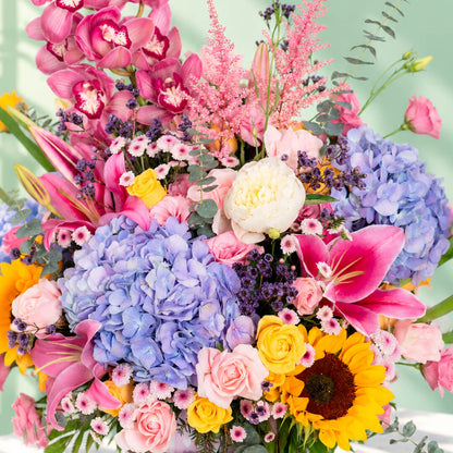 Pink round flower box featuring 50–60 stems of assorted hydrangeas, peonies, sunflowers, tulips, roses, lilies, cymbidium orchids and eucalyptus greenery.
