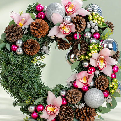 Pink orchid Christmas wreath with evergreen foliage, pine cones, berries, and silver ornaments for festive décor.