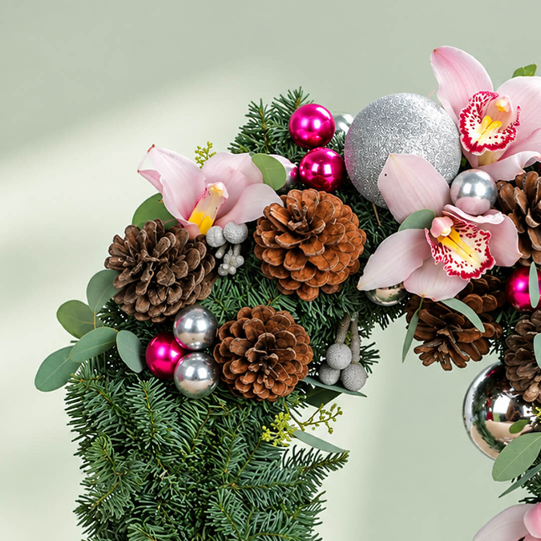 Pink orchid Christmas wreath with evergreen foliage, pine cones, berries, and silver ornaments for festive décor.