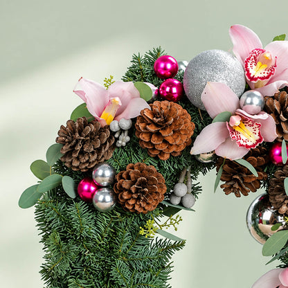 Pink orchid Christmas wreath with evergreen foliage, pine cones, berries, and silver ornaments for festive décor.