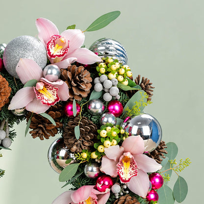 Pink orchid Christmas wreath with evergreen foliage, pine cones, berries, and silver ornaments for festive décor.