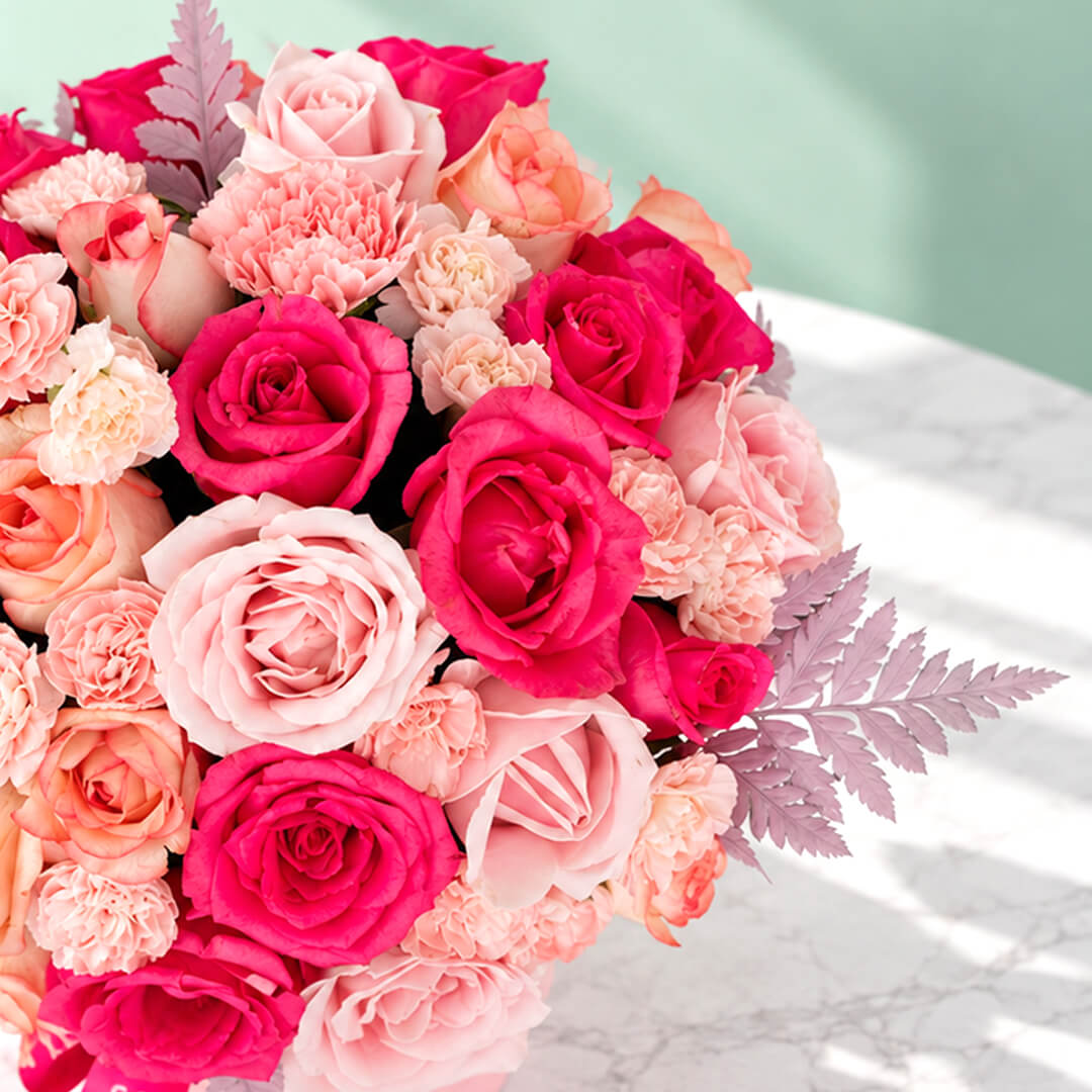 Round pink flower box filled with pink roses and carnations, accented with fresh green leaves.