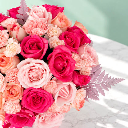 Round pink flower box filled with pink roses and carnations, accented with fresh green leaves.