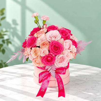 Round pink flower box filled with pink roses and carnations, accented with fresh green leaves.