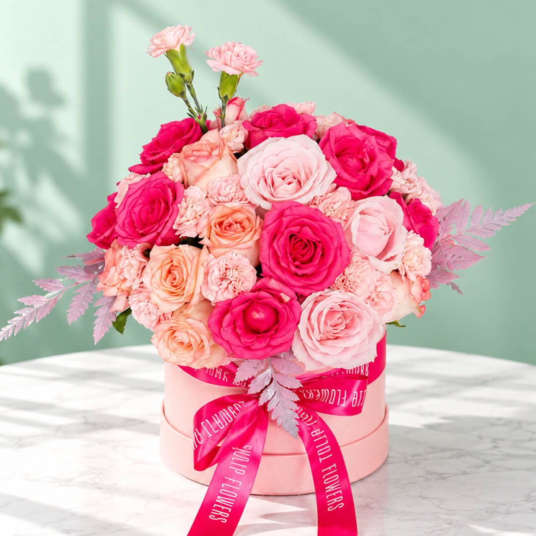 Round pink flower box filled with pink roses and carnations, accented with fresh green leaves.