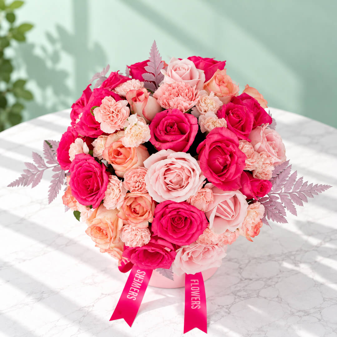 Round pink flower box filled with pink roses and carnations, accented with fresh green leaves.