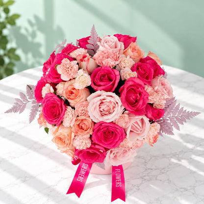 Round pink flower box filled with pink roses and carnations, accented with fresh green leaves.