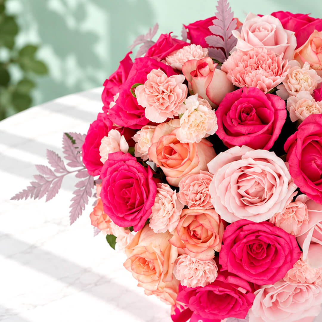 Round pink flower box filled with pink roses and carnations, accented with fresh green leaves.
