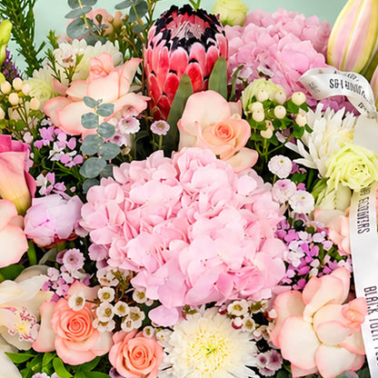 Assorted flowers including roses, hydrangea, calla lily, cymbidium, and Queen Protea arranged in a rectangular basket