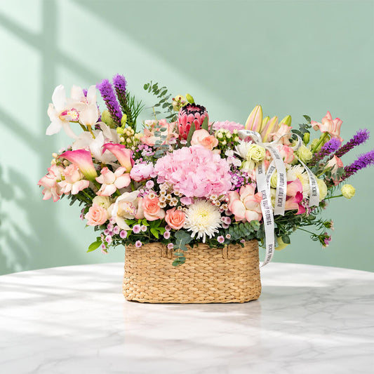 Assorted flowers including roses, hydrangea, calla lily, cymbidium, and Queen Protea arranged in a rectangular basket