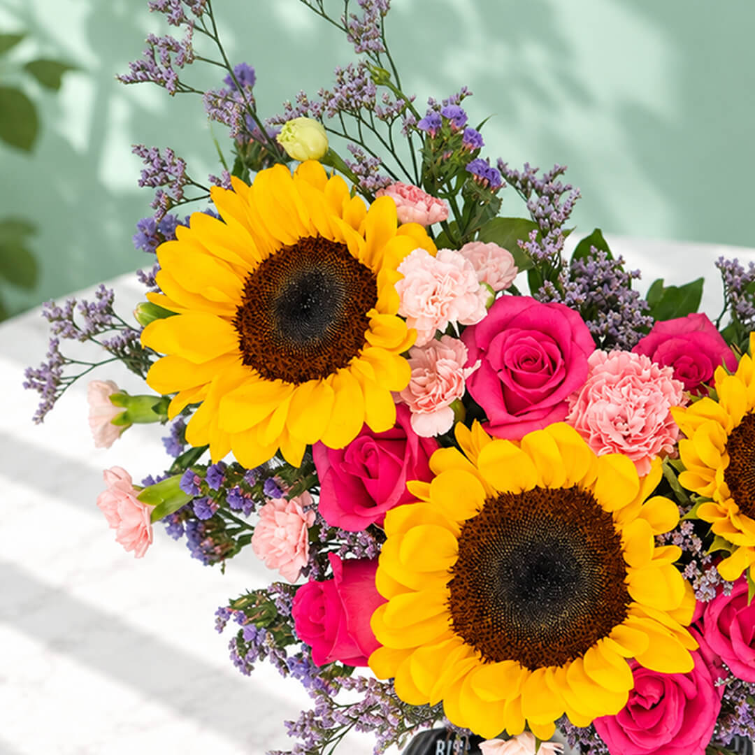 Pink round flower box featuring sunflowers, spray carnations, statice and green leaves in assorted colours.