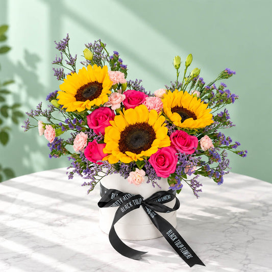 Pink round flower box featuring sunflowers, spray carnations, statice and green leaves in assorted colours.