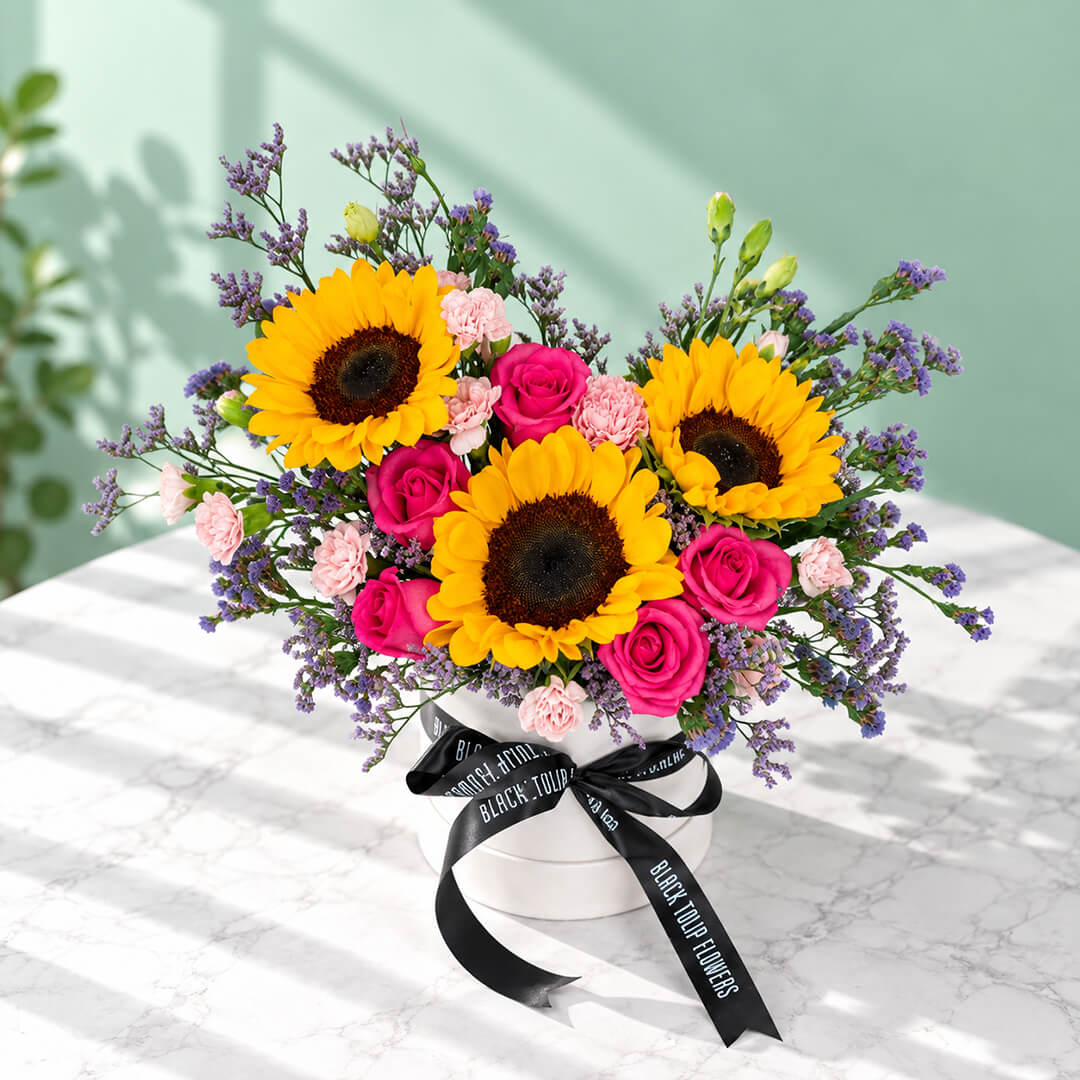 Pink round flower box featuring sunflowers, spray carnations, statice and green leaves in assorted colours.