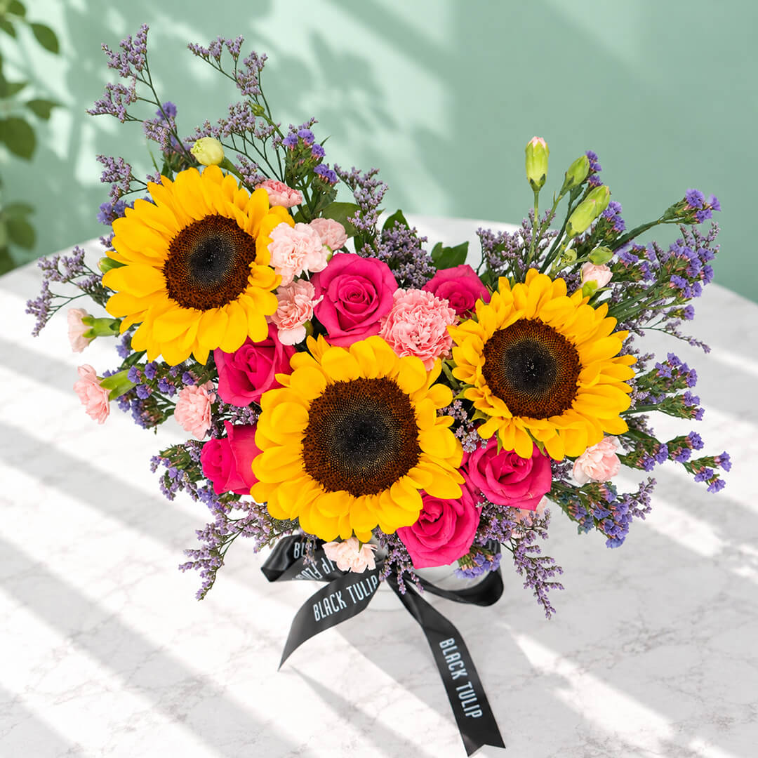Pink round flower box featuring sunflowers, spray carnations, statice and green leaves in assorted colours.