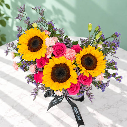 Pink round flower box featuring sunflowers, spray carnations, statice and green leaves in assorted colours.