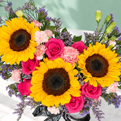 Pink round flower box featuring sunflowers, spray carnations, statice and green leaves in assorted colours.