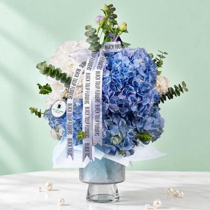 Blue hydrangea vase arrangement with fresh greenery and white ribbon accents
