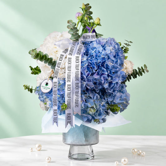 Blue hydrangea vase arrangement with fresh greenery and white ribbon accents
